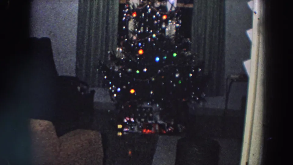 1958 Italy home-movie frame showing a lit Christmas tree with ornaments, curtains, chairs, and a doorway edge in a private living room.