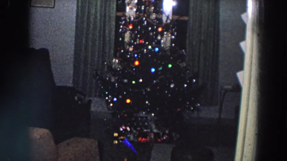 1958 Italy archival home-movie frame of a Christmas tree with multicolored lights, ornaments, curtains, furniture, and partial doorway framing.