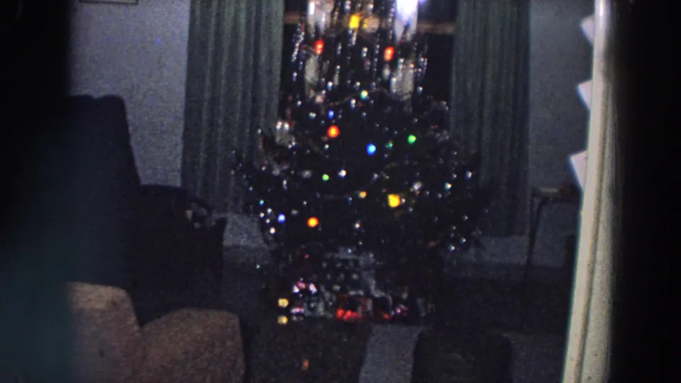 1958 Italy 8mm frame showing a large Christmas tree with bright colored bulbs, tinsel-like reflections, gifts, curtains, and living-room seating.