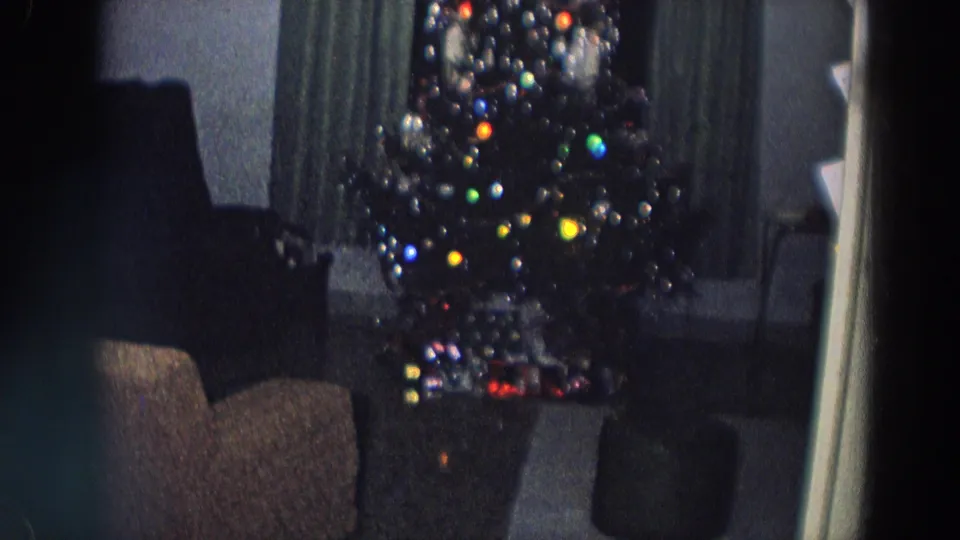 1958 Italy home-movie frame of a dim living room with a decorated Christmas tree, glowing bulbs, curtains, chairs, and gifts at the base.