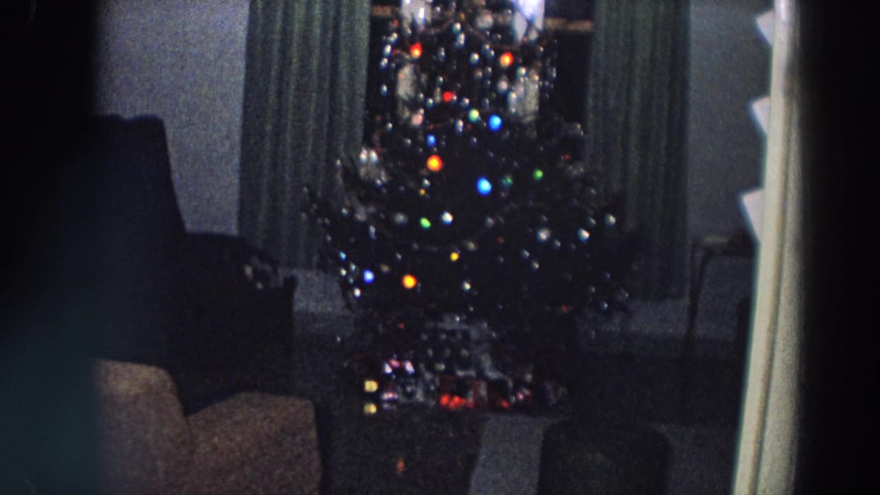 1958 Italy home-movie frame showing a lit Christmas tree before dark curtains in a dim private living room.