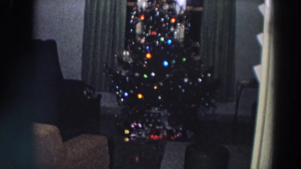 1958 Italy home-movie frame showing a lit Christmas tree before dark curtains in a dim private living room.
