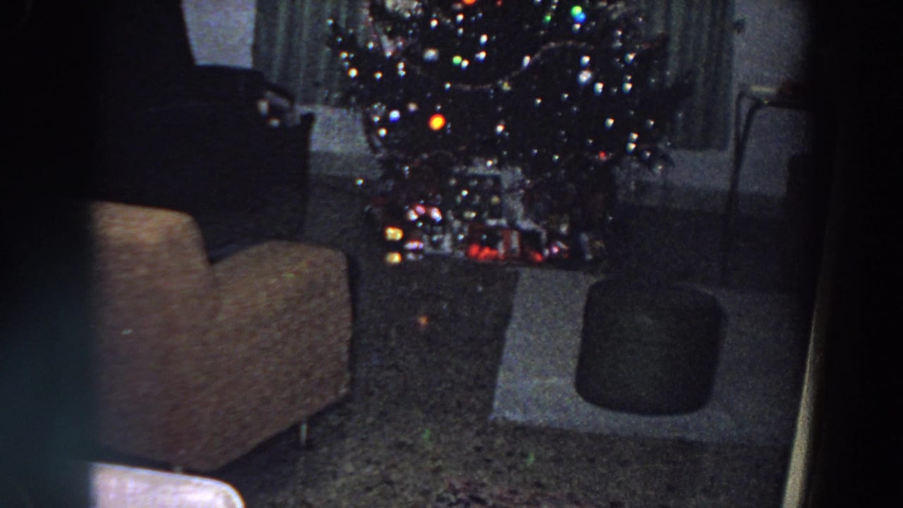 1958 Italy 8mm frame showing a dim living room with a lit Christmas tree, wrapped gifts, armchair, ottoman, and curtains.