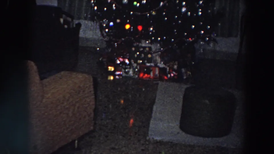 1958 Italy 8mm view of the lower half of a decorated Christmas tree with gifts, ornaments, armchair, and ottoman visible.