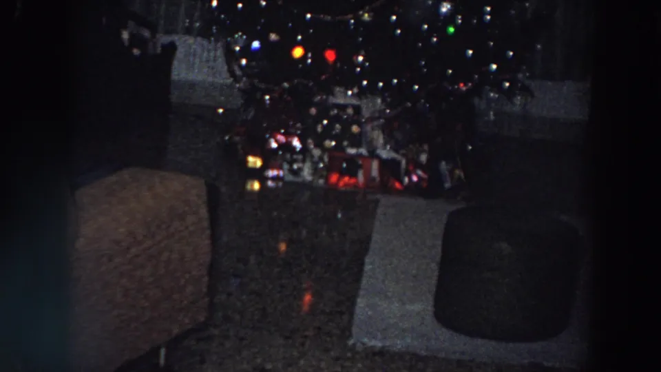 1958 Italy frame of a dim domestic Christmas scene with tree lights, gifts, an armchair, ottoman, curtains, and reflective floor patches.