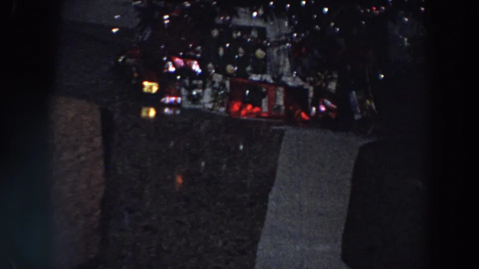 Grainy 8mm frame from 1958 Italy showing the tree lights and wrapped presents in a dim living room.