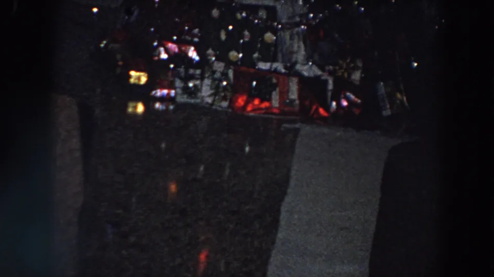 1958 Italian home-movie frame with colored Christmas lights, wrapped presents, and surrounding dark furniture.