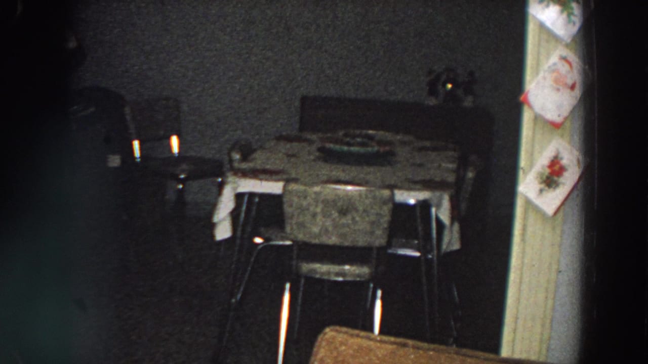 Dim dining room with table, chairs, hanging Christmas cards, and a partially visible person at left.