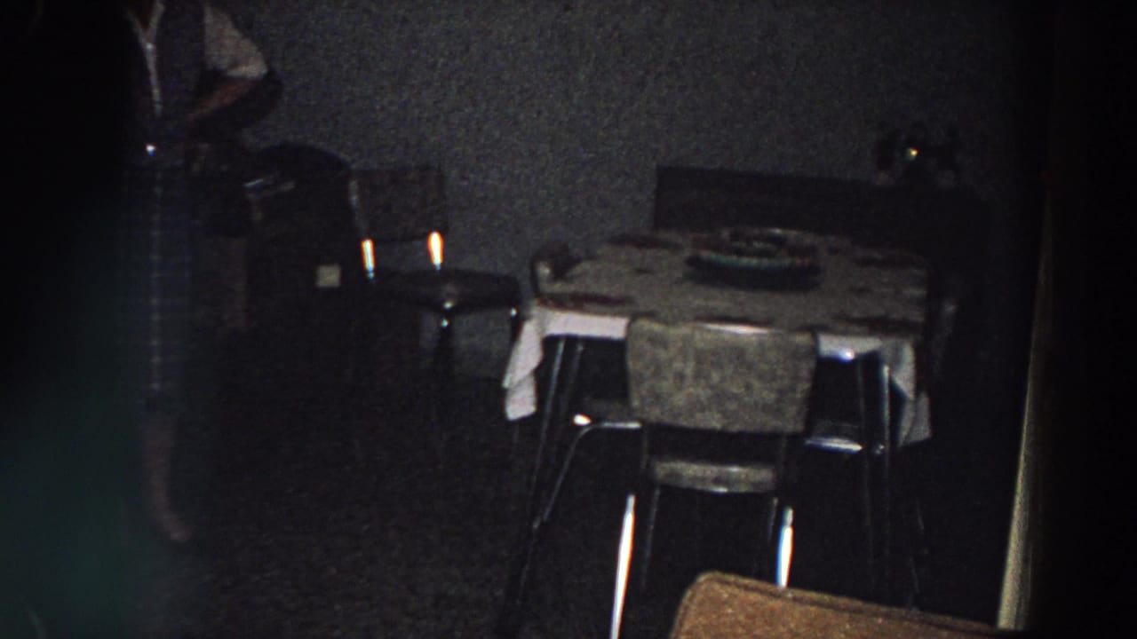 1958 Italy interior frame with a dining table, chairs, dark wall, red light fixture, and a partially visible person moving through the room.