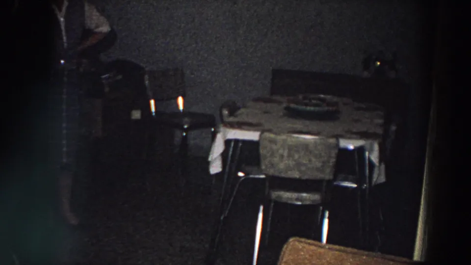 1958 Italy interior frame with a dining table, chairs, dark wall, red light fixture, and a partially visible person moving through the room.