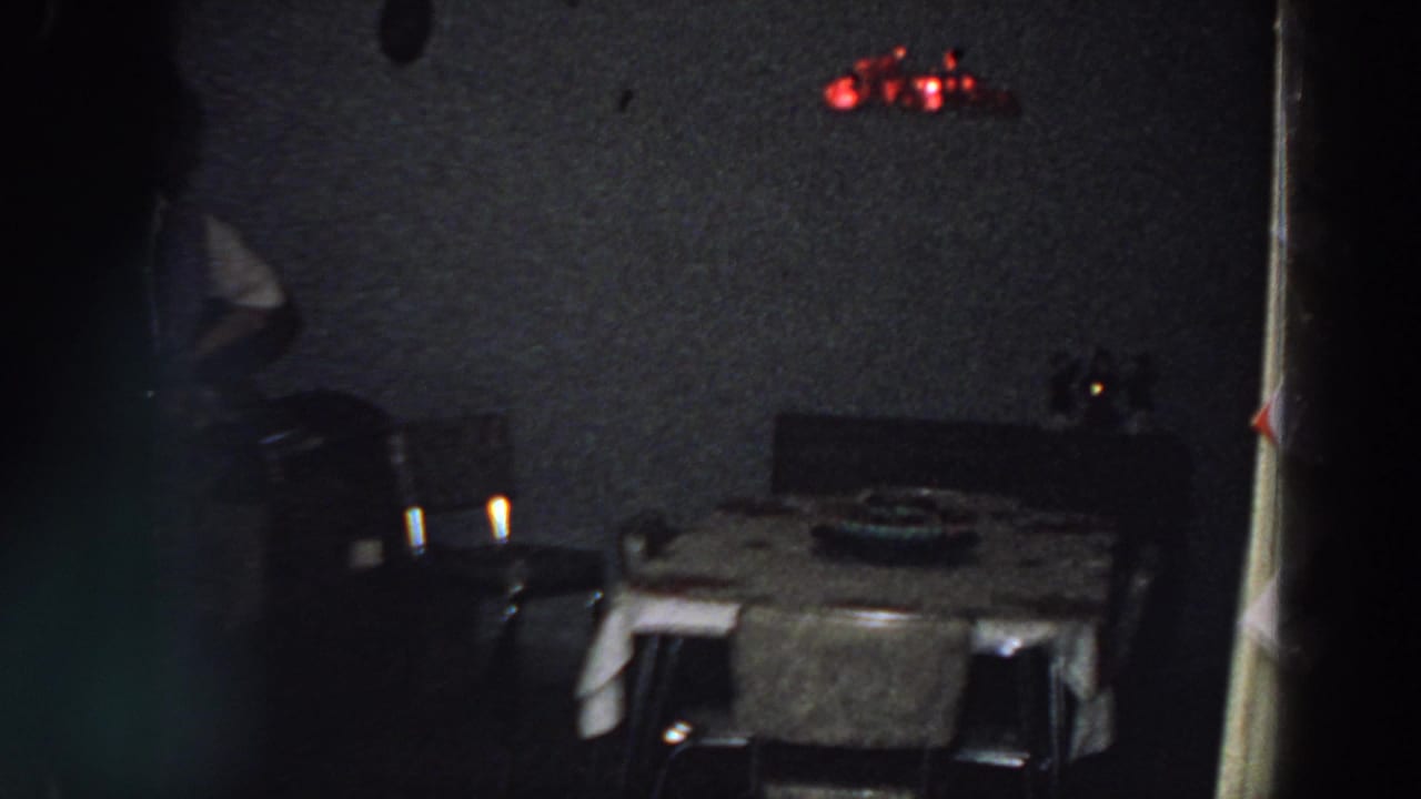 1958 Italy home interior showing a dining table under dim lighting, chairs, red wall light, doorway edge, and a person partly entering frame.