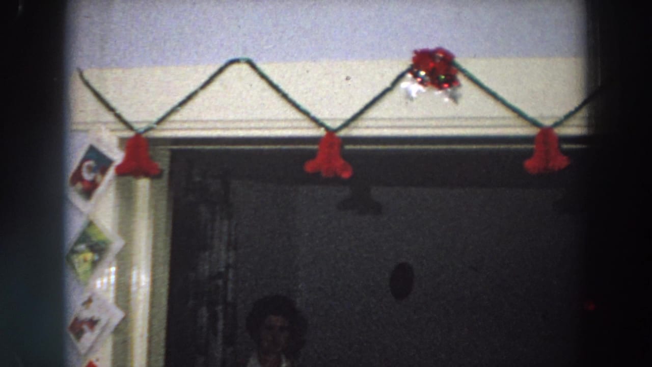 1958 Italy holiday interior with red garland, taped Christmas cards, and a faint figure in the dark doorway beneath the decorations.