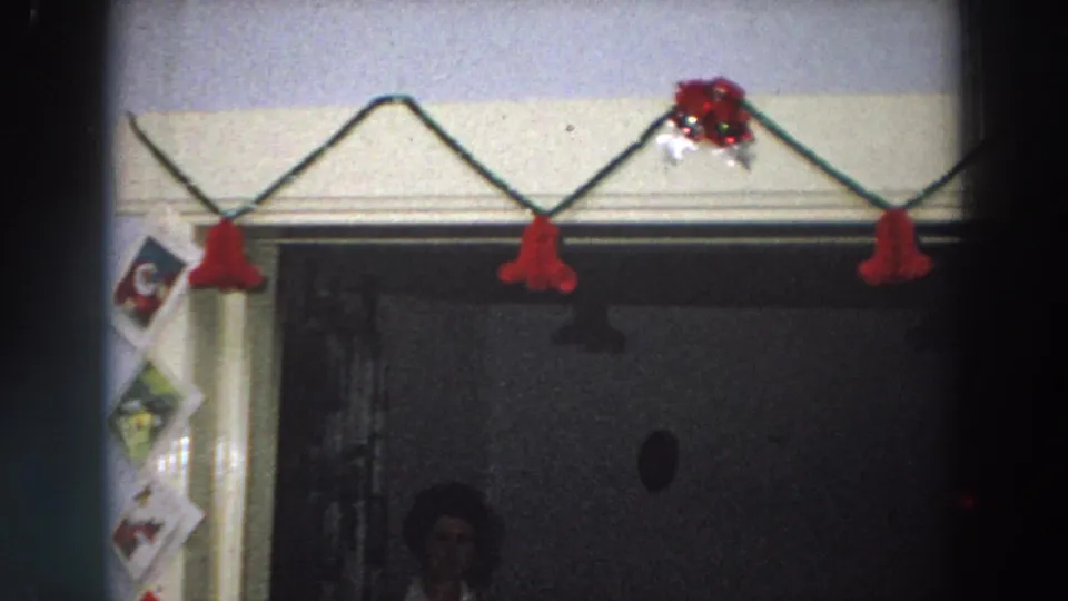 1958 Italy holiday interior with red garland, taped Christmas cards, and a faint figure in the dark doorway beneath the decorations.