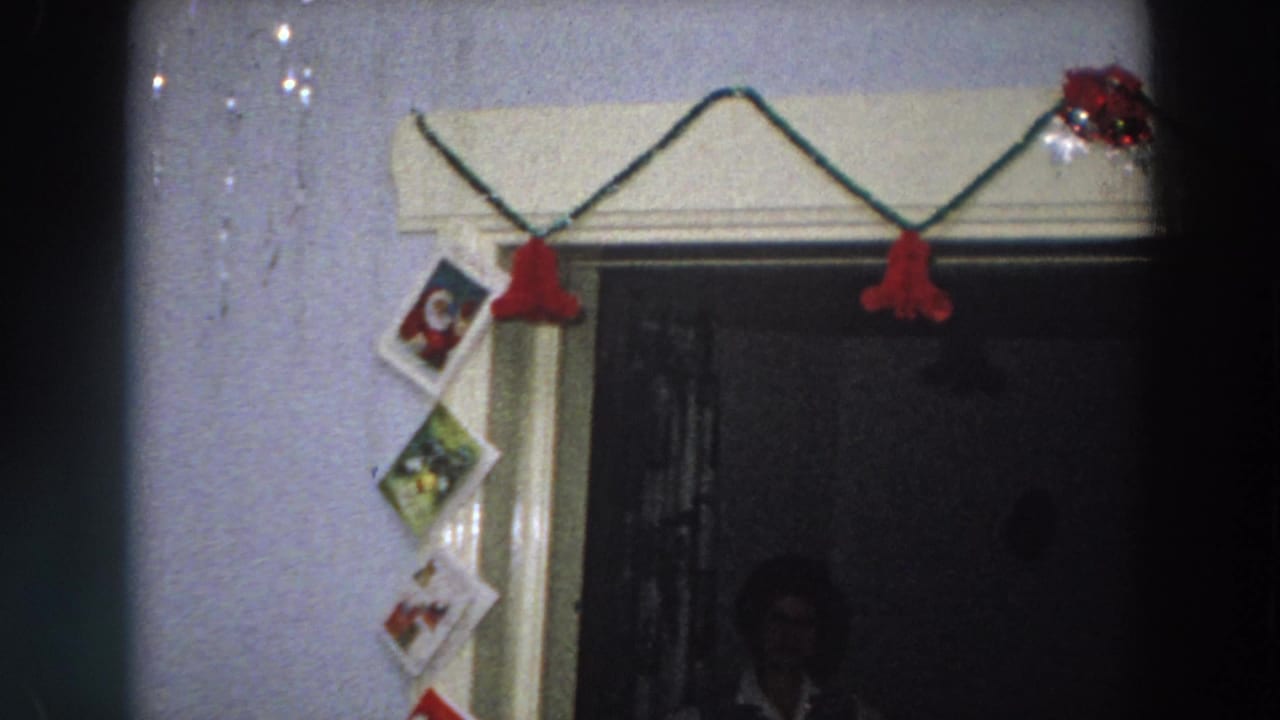 1958 Italy 8mm view of Christmas cards taped beside a decorated doorway, with red bell garland and dark interior space visible.
