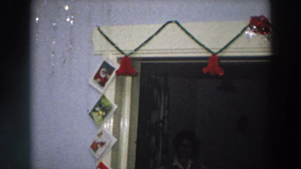 1958 Italy 8mm view of Christmas cards taped beside a decorated doorway, with red bell garland and dark interior space visible.
