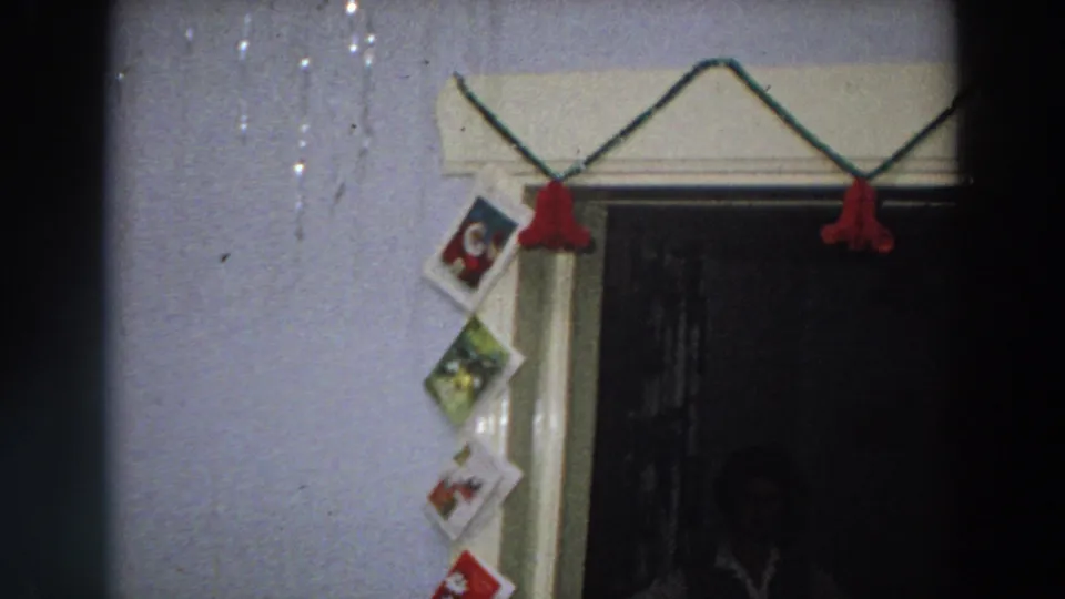 1958 Italy home-movie frame showing red Christmas garland over a doorway and greeting cards taped vertically along the wall.
