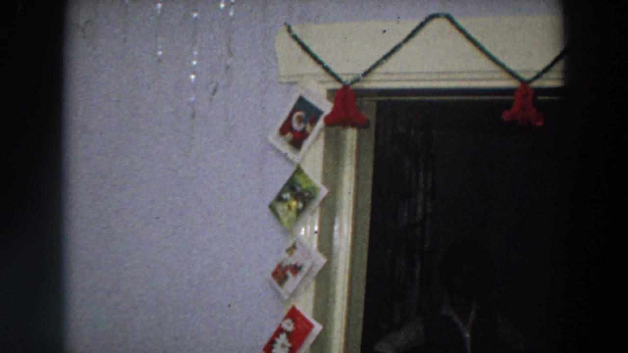 1958 Italy home-movie frame showing Christmas cards strung along a pale wall beside a dark doorway with red tassels above.