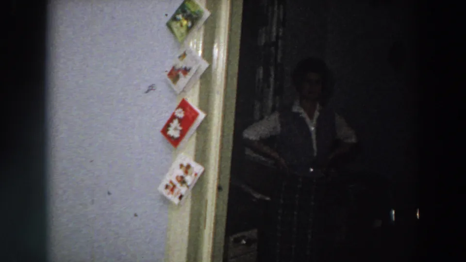 1958 Italy frame with a person in a dark vest and light shirt standing in a dim doorway beside a vertical display of Christmas cards.