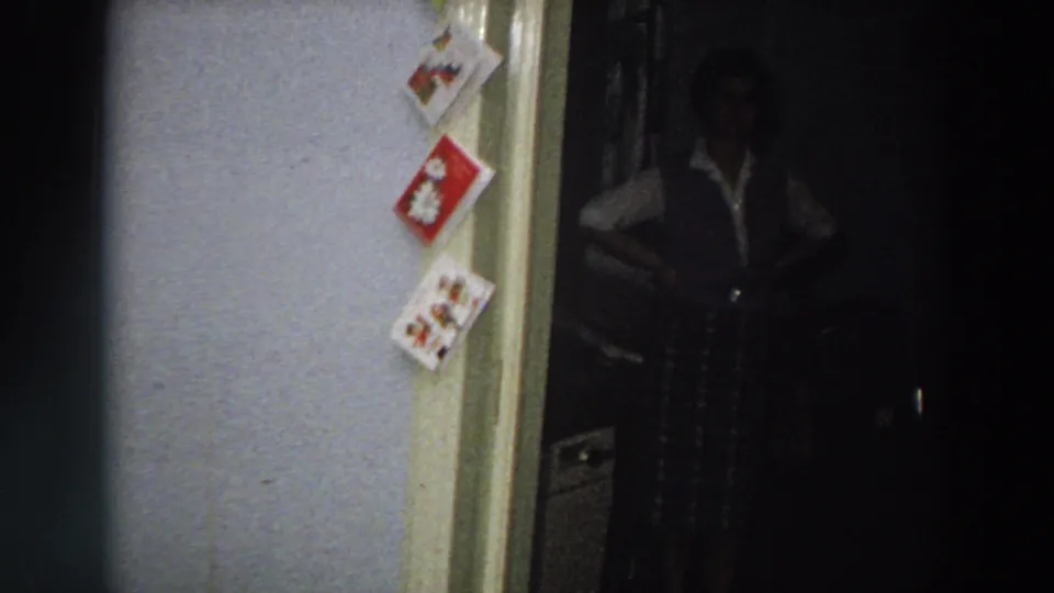 1958 Italy home-movie frame showing a dim private bedroom doorway, pinned Christmas cards, and a woman standing partly in shadow.