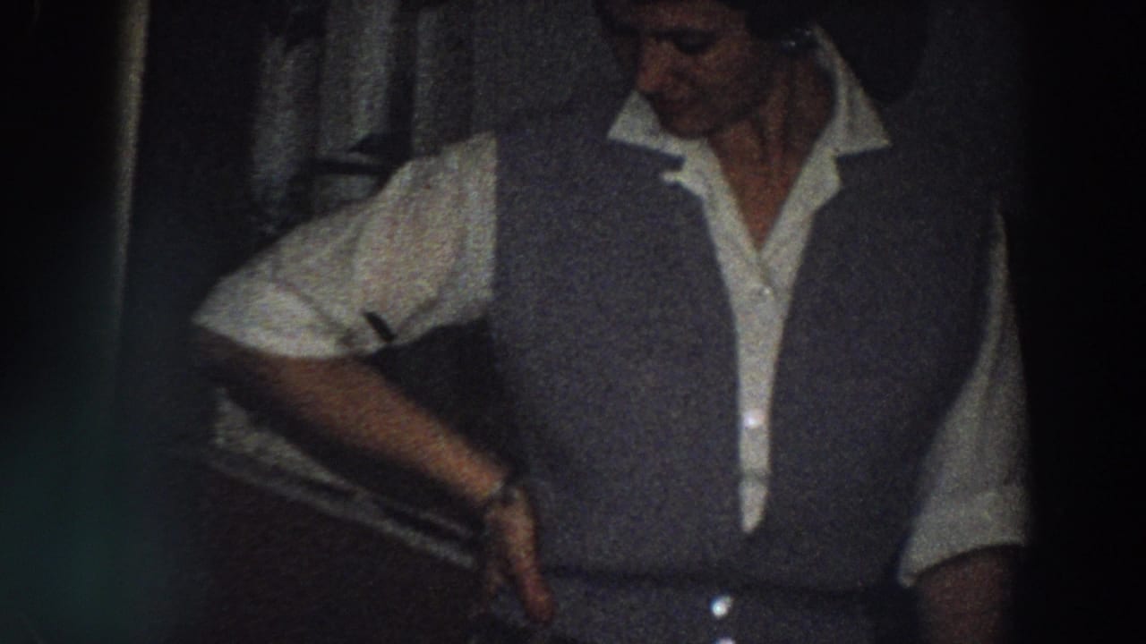 1958 Italy home-movie frame showing an adult woman posing in a grey vest, white shirt, and plaid skirt inside a dark bedroom.