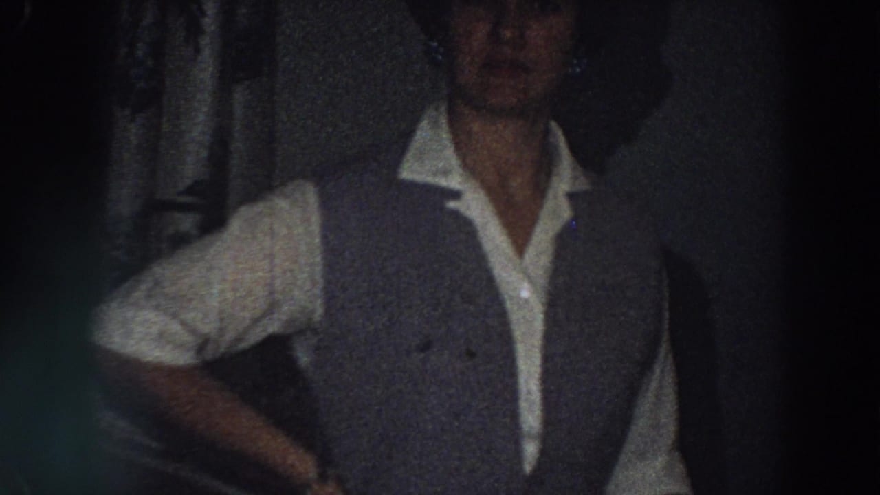 1958 Italy home-movie frame of an adult woman facing the camera in a dark private bedroom, with her grey vest and white shirt visible.