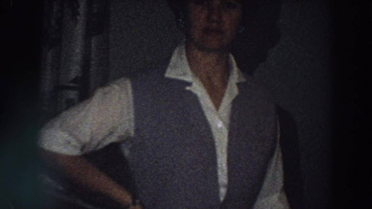 Adult woman in a vest stands indoors beside a dining area.