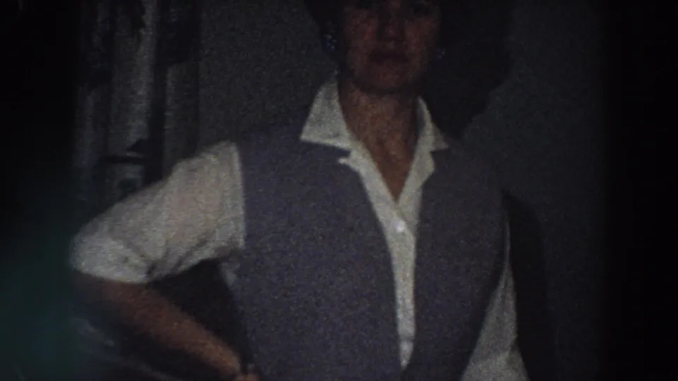 In a 1958 Italian home interior, an adult woman in a white shirt and dark vest stands near a dining table.