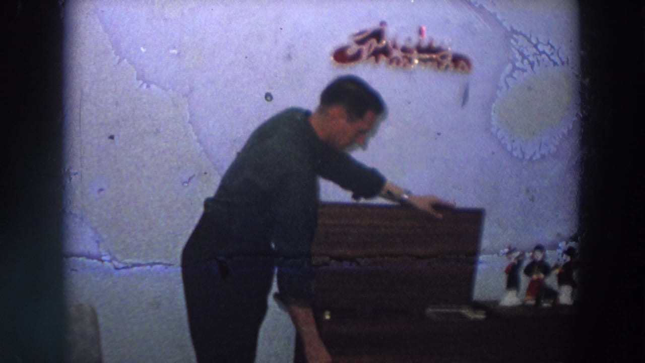 In a 1958 Italy home interior, a man bends over a wooden storage box beneath a decorative Christmas sign.