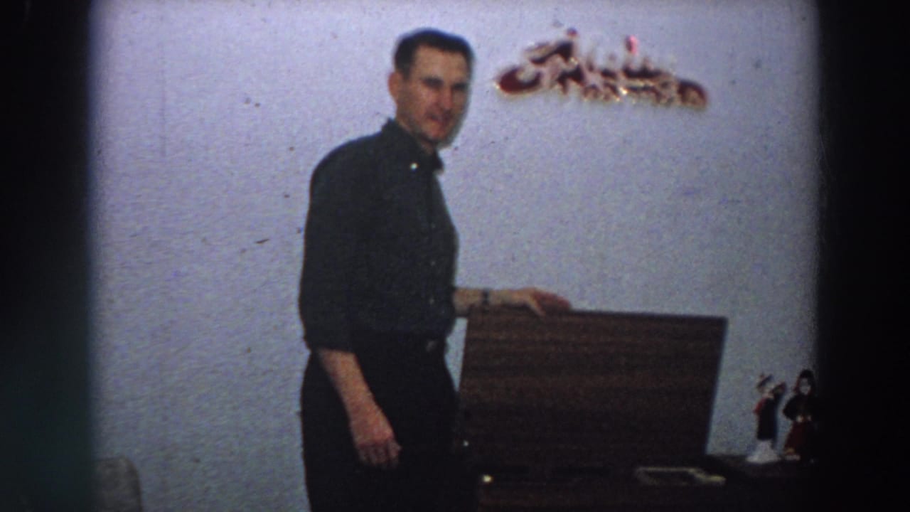 The man stands beside the wooden box in a modest 1958 Italy interior, turning toward the camera with a slight smile.