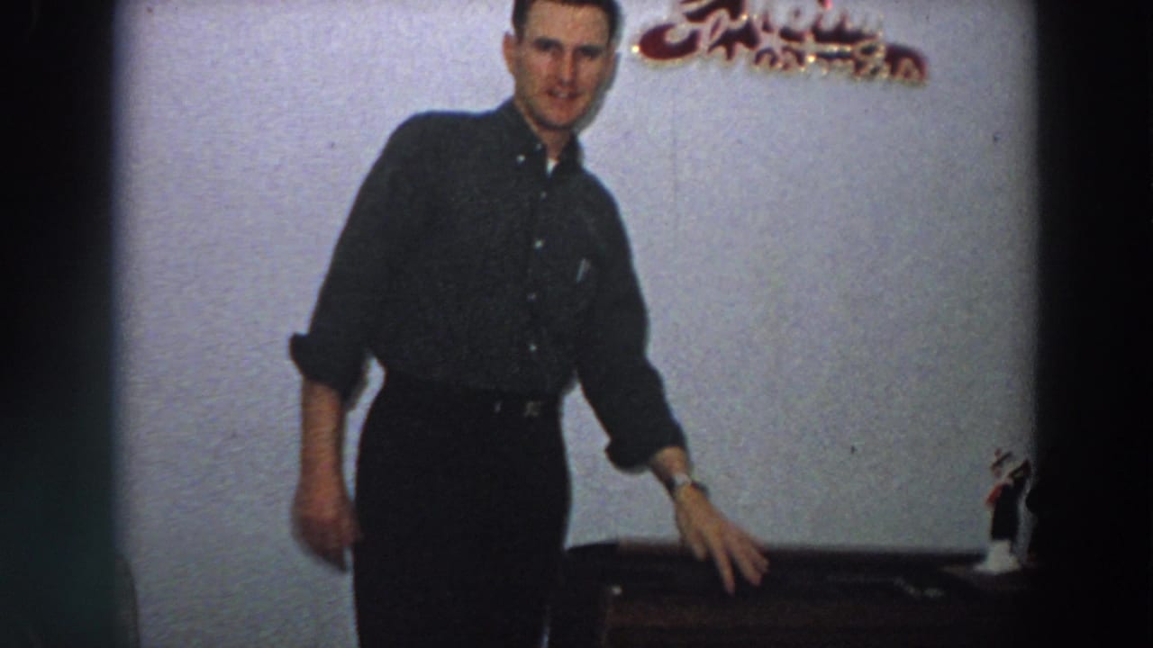 A man in a dark shirt stands near the wooden box, with a Christmas decoration and figurine visible in the home interior.