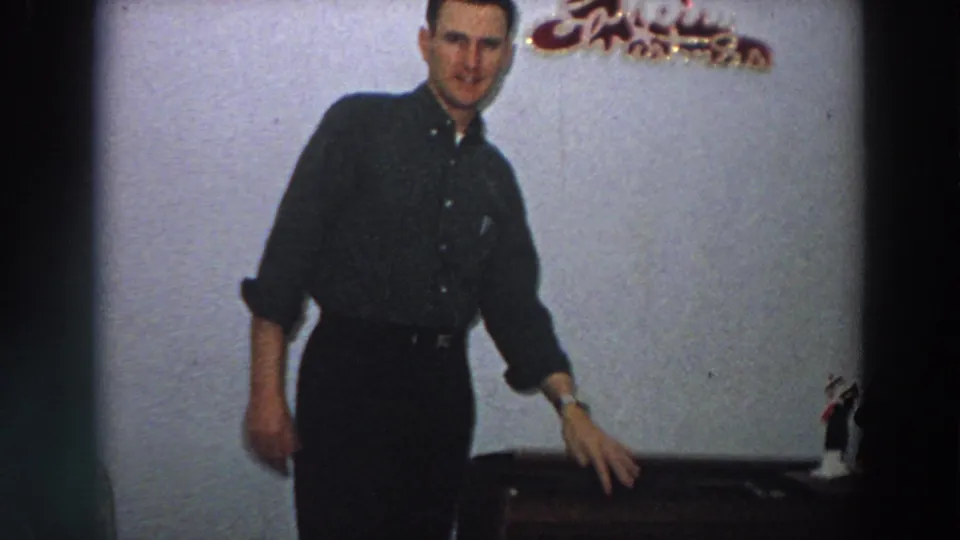 A man in a dark shirt stands near the wooden box, with a Christmas decoration and figurine visible in the home interior.