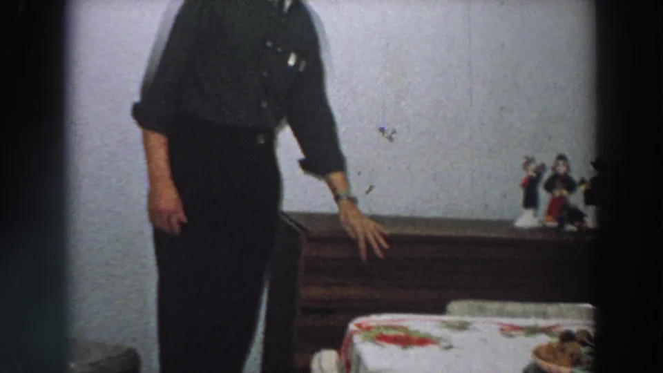 The man rests a hand near the wooden box while a bed edge and small figurines appear in the 1958 Italy home interior.
