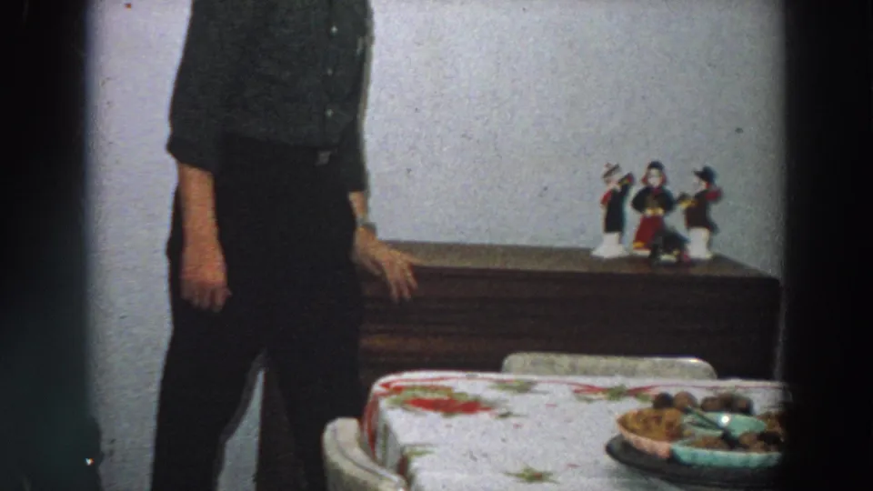 In a 1958 Italian home interior, an adult man stands partly cropped beside a floral-covered dining table and wooden dresser.
