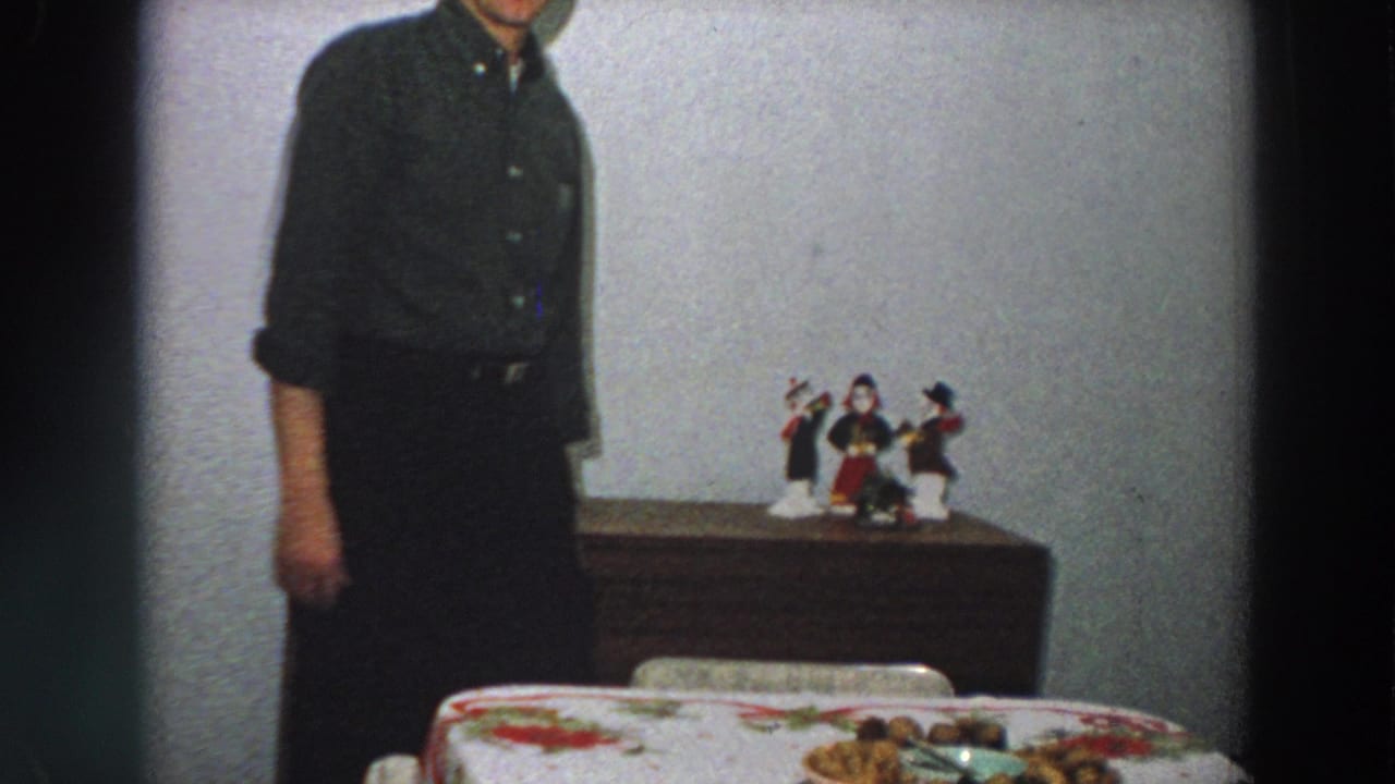 An adult man in dark clothing stands in a modest 1958 Italian interior near a dining table and dresser with figurines.