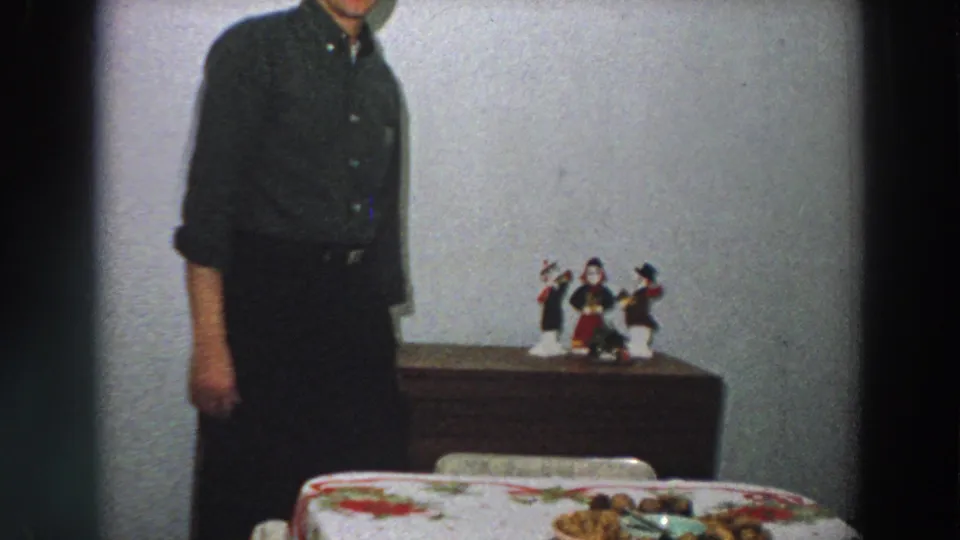 An adult man in dark clothing stands in a modest 1958 Italian interior near a dining table and dresser with figurines.