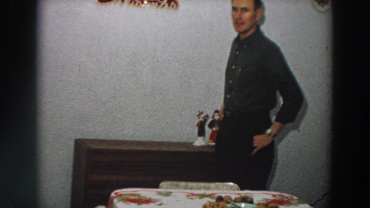 The man faces toward the camera in a 1958 Italian home scene, standing between a dining table and a wooden dresser.