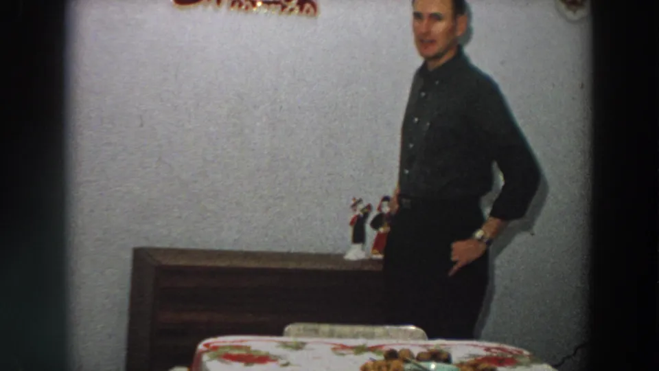 The man faces toward the camera in a 1958 Italian home scene, standing between a dining table and a wooden dresser.