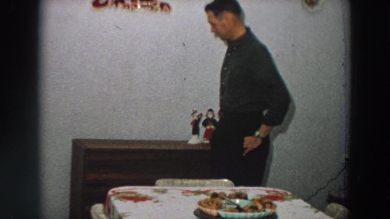 Man in dark clothing stands beside a dining table with a floral cloth and plate of food.