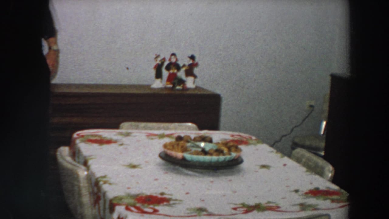 The room is briefly shown without the man's face, emphasizing the floral tablecloth, plate of food, sideboard, and figurines.
