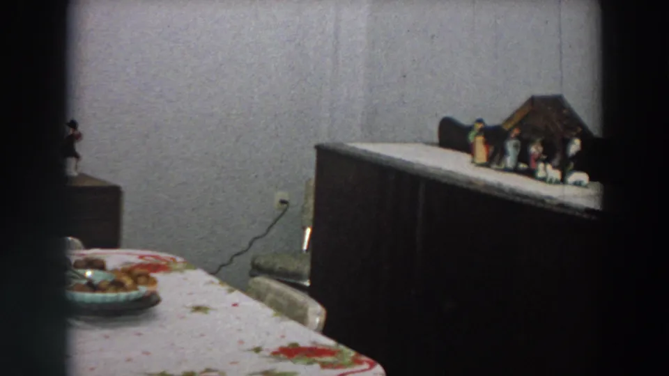 1958 Italian home-movie frame showing a dim domestic room with a floral tablecloth, dark cabinet, and small nativity display.