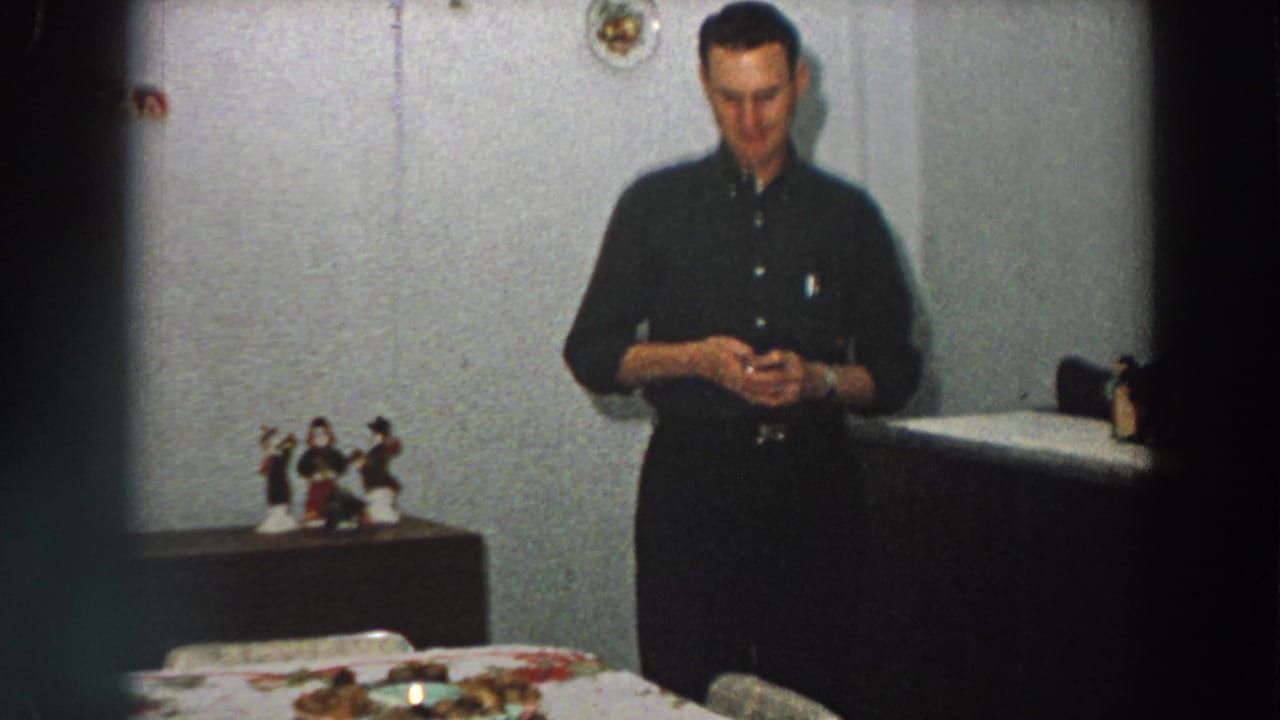 1958 Italian home-movie frame showing an adult man in a dark shirt standing near a table, cabinet, and small religious figurines.