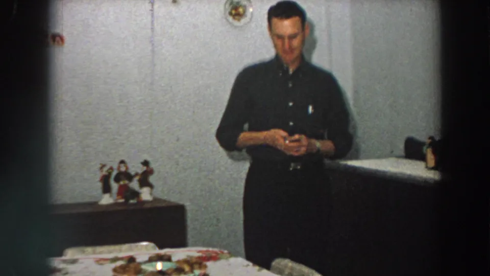 1958 Italian home-movie frame showing an adult man in a dark shirt standing near a table, cabinet, and small religious figurines.