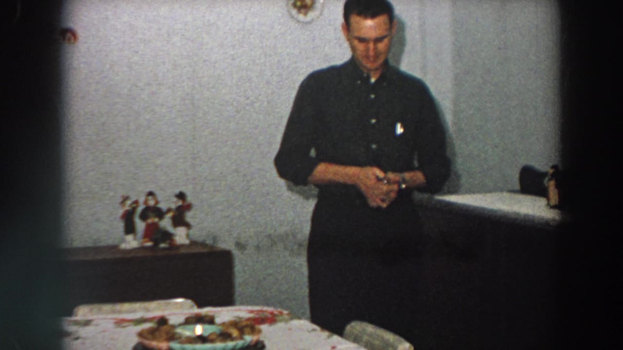In 1958 Italy, a man in a dark shirt stands indoors, looking down at a small box near a table with figurines and a patterned cloth.
