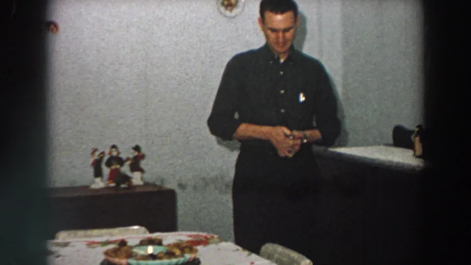 In 1958 Italy, a man in a dark shirt stands indoors, looking down at a small box near a table with figurines and a patterned cloth.