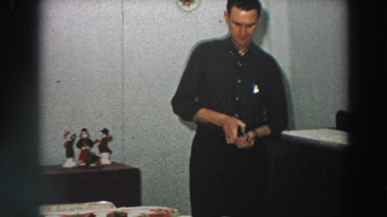 The man continues opening or examining the small box in a plain Italian home interior, with figurines and a table visible nearby.