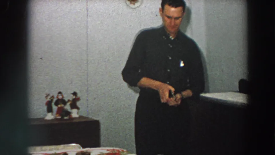 The man continues opening or examining the small box in a plain Italian home interior, with figurines and a table visible nearby.