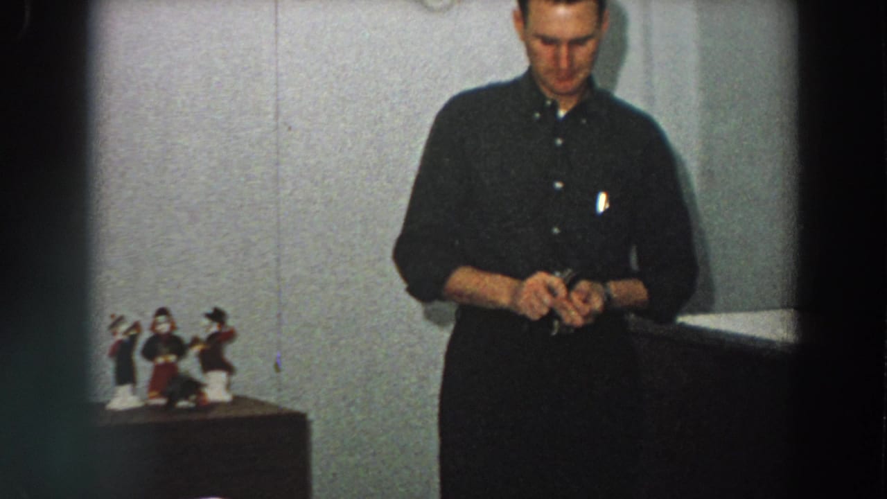 A closer home-movie frame shows the man focused on the small box, with tabletop figurines and a plain wall behind him.