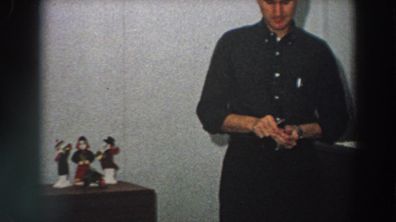 Adult man in a dark shirt stands indoors beside small tabletop figurines.