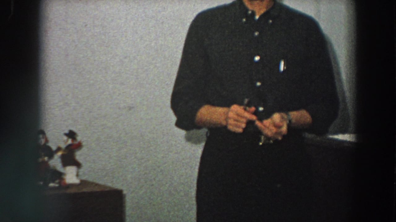 An adult man in dark clothing handles a small item indoors, with decorative figurines visible at the side of the frame.