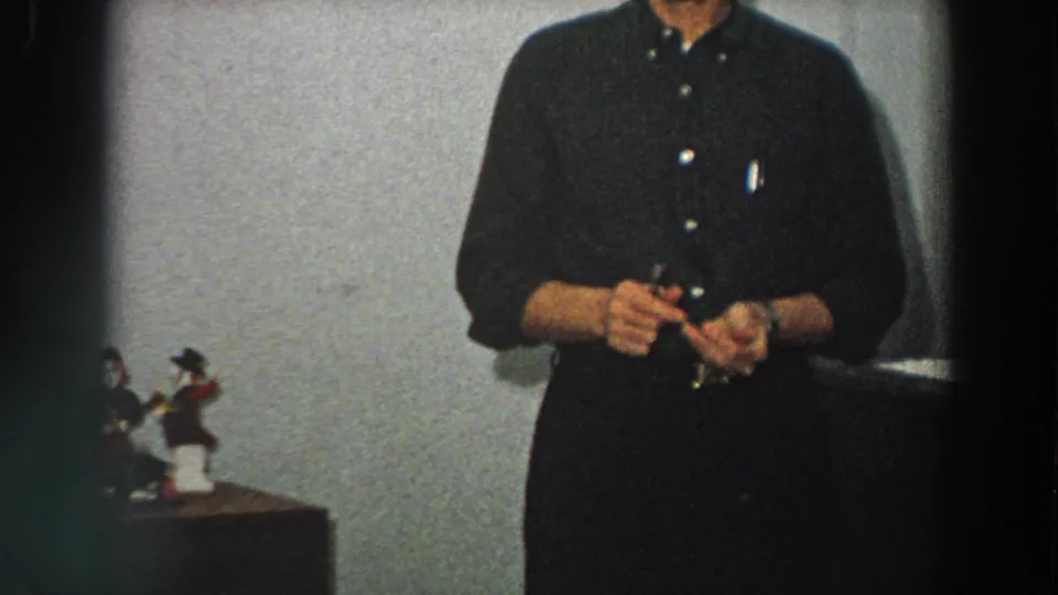 An adult man in dark clothing handles a small item indoors, with decorative figurines visible at the side of the frame.